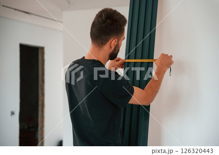 Taking measurements. Plumber worker installing heating radiator in empty room of a newly built house 126320443