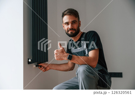 Sitting and showing thumb up. Plumber worker installing heating radiator in empty room of a newly built house 126320561