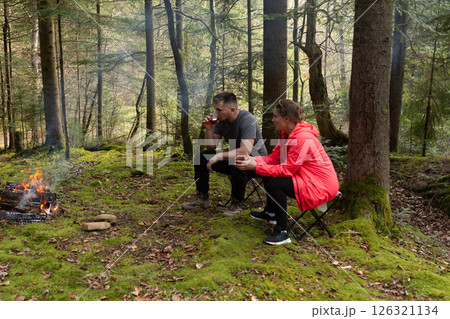 Couple enjoys cozy moments by a warm fire in a lush forest during a serene afternoon retreat with steaming drinks and nature's tranquility 126321134