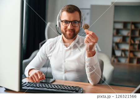 Holding bitcoin in the hand. Man in formal clothes is working in the modern office. Using computer Holding bitcoin in the hand. Man in formal clothes is working in the modern office. Using computer 126321566