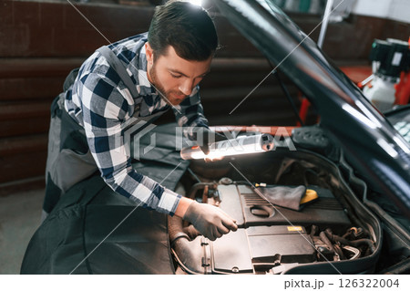 Under the hood. Holding lighting equipment and fixing the issues of the car. Man in uniform is working in the auto salon Under the hood. Holding lighting equipment and fixing the issues of the car. Man in uniform is working in the auto salon 126322004