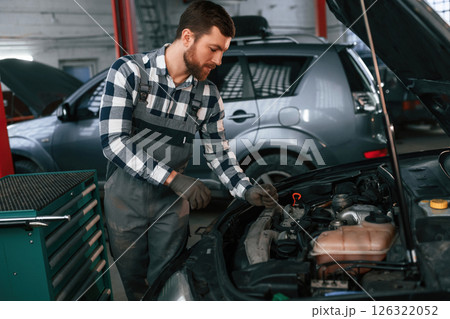 Mechanic is using a tool to repair the car under the hood. Man in uniform is working in the auto salon 126322052
