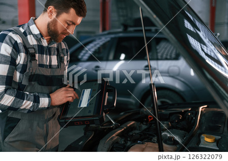 Standing near the car with hood opened. Holding tablet. Man in uniform is working in the auto salon Standing near the car with hood opened. Holding tablet. Man in uniform is working in the auto salon 126322079