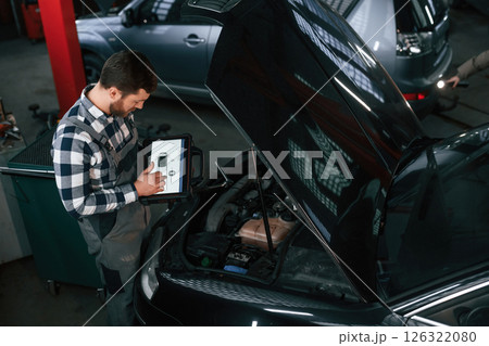 Standing near the car with hood opened. Holding tablet. Man in uniform is working in the auto salon Standing near the car with hood opened. Holding tablet. Man in uniform is working in the auto salon 126322080