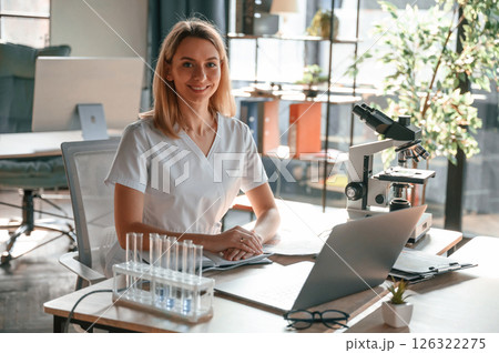 Smiling, cheerful mood. Young female doctor in coat is sitting in the clinic and working 126322275