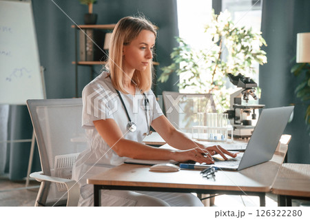 Using laptop. Young female doctor in coat is sitting in the clinic and working 126322280