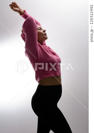 Pregnant woman in pink sweater and black leggings, arms raised, against a light background. Pregnant woman in pink sweater and black leggings, arms raised, against a light background. 126322492