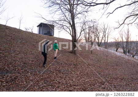Two individuals exploring a leaf covered hillside during early evening in a tranquil landscape with bare trees and a small shelter in the background 126322882