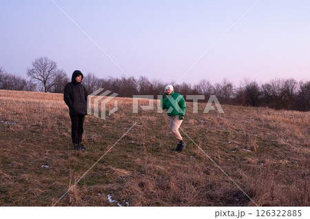 Two friends enjoying a playful moment on a grassy hill at sunset, surrounded by trees, embracing the tranquility of nature in late winter Two friends enjoying a playful moment on a grassy hill at sunset, surrounded by trees, embracing the tranquility of nature in late winter 126322885