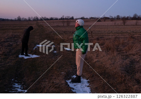 Two figures stand in a rural landscape at dusk, one dressed warmly in green while the other quietly bows, capturing moments of solitude and reflection in nature 126322887
