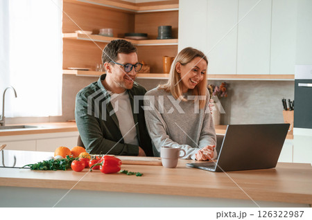Sitting, using laptop. Young couple is on the kitchen together Sitting, using laptop. Young couple is on the kitchen together 126322987