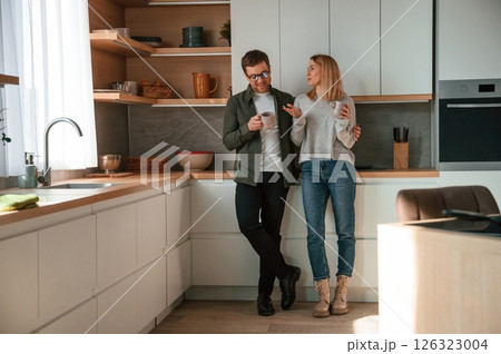 Standing, talking, holding cups with drink. Young couple is on the kitchen together 126323004