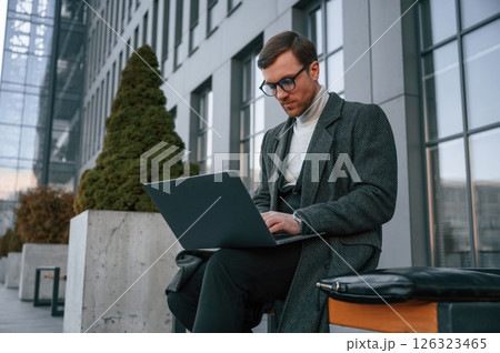 Sitting on the bench with laptop. Handsome man in formal clothes is outdoor near the business building 126323465
