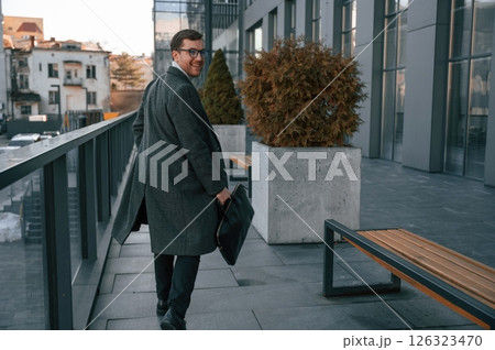Holding the bag. Handsome man in formal clothes is outdoor near the business building 126323470