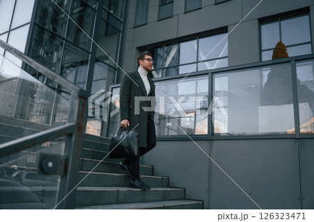 Holding the bag. Handsome man in formal clothes is outdoor near the business building Holding the bag. Handsome man in formal clothes is outdoor near the business building 126323471