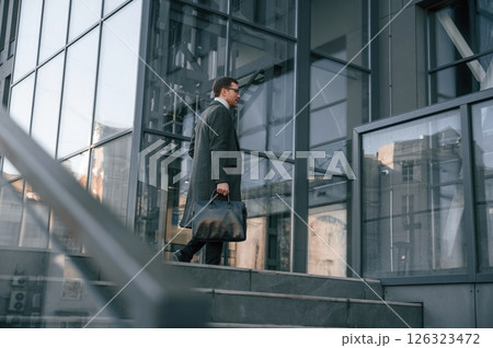 Holding the bag. Handsome man in formal clothes is outdoor near the business building Holding the bag. Handsome man in formal clothes is outdoor near the business building 126323472