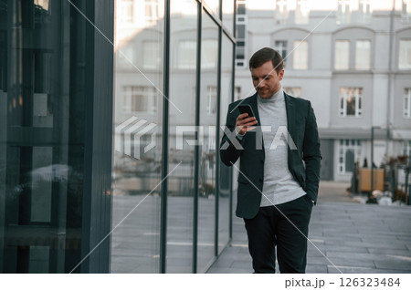 Using social media by smartphone. Handsome man in formal clothes is outdoor near the business building Using social media by smartphone. Handsome man in formal clothes is outdoor near the business building 126323484