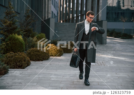 Checking the time. Handsome man in formal clothes is outdoor near the business building Checking the time. Handsome man in formal clothes is outdoor near the business building 126323759