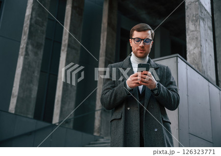 Black colored smartphone in hands. Handsome man in formal clothes is outdoor near the business building Black colored smartphone in hands. Handsome man in formal clothes is outdoor near the business building 126323771