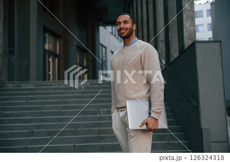 Positive handsome black man is outdoors near the business building holding laptop 126324318