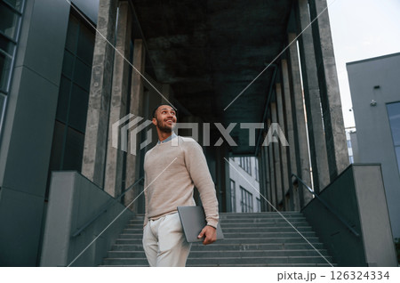 Silver colored laptop in hands. Handsome black man is outdoors near the business building 126324334