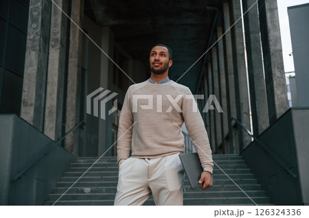 Silver colored laptop in hands. Handsome black man is outdoors near the business building 126324336