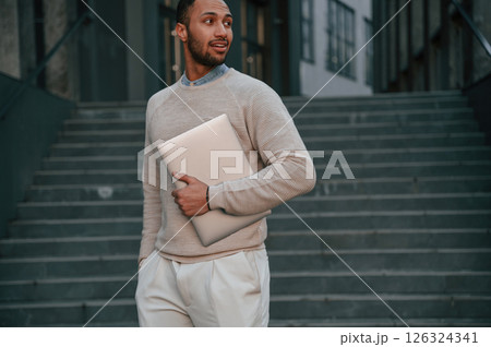 Holding laptop in hand. Handsome black man is outdoors near the business building 126324341