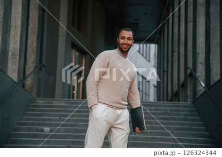 Holding laptop in hand. Handsome black man is outdoors near the business building Holding laptop in hand. Handsome black man is outdoors near the business building 126324344