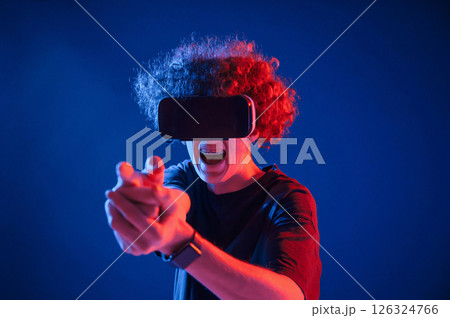 Pistol gesture by hands. In VR glasses. Young man with curly hair is indoors illuminated by neon lighting Pistol gesture by hands. In VR glasses. Young man with curly hair is indoors illuminated by neon lighting 126324766