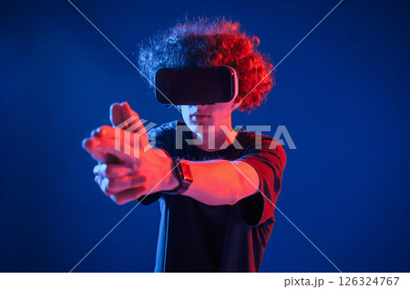 Pistol gesture by hands. In VR glasses. Young man with curly hair is indoors illuminated by neon lighting 126324767
