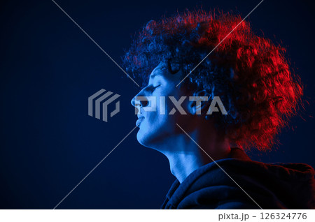 View from the side. Young man with curly hair is indoors illuminated by neon lighting View from the side. Young man with curly hair is indoors illuminated by neon lighting 126324776