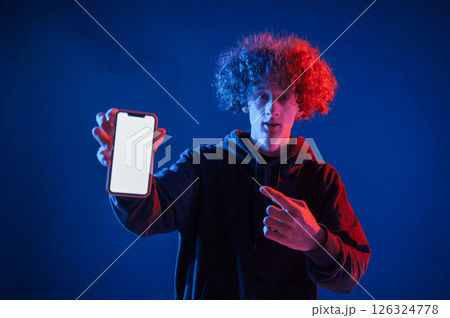 Holding smartphone and showing it to the camera. Young man with curly hair is indoors illuminated by neon lighting Holding smartphone and showing it to the camera. Young man with curly hair is indoors illuminated by neon lighting 126324778