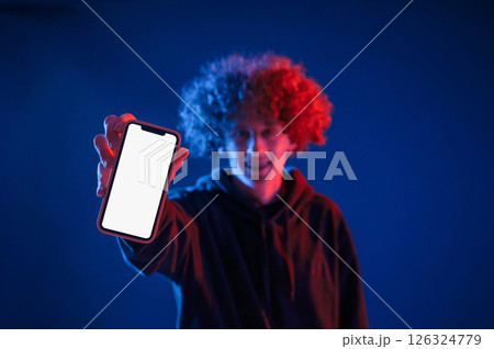 Holding smartphone and showing it to the camera. Young man with curly hair is indoors illuminated by neon lighting 126324779