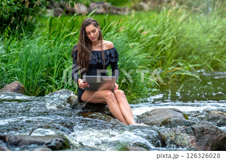 woman freelancer sitting on a rock in the river and using a laptop, travel or vacation work concept 126326028