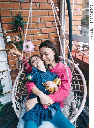 Mother and daughter hugging in a wicker garden swing surrounded by flowers. The concept of a happy childhood and loving caring parents Mother and daughter hugging in a wicker garden swing surrounded by flowers. The concept of a happy childhood and loving caring parents 126326567