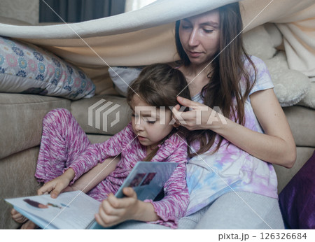 Mother and daughter sit in a hut made of a blanket and read a book. Pleasant time with children, quiet time before bed. Mother and daughter sit in a hut made of a blanket and read a book. Pleasant time with children, quiet time before bed. 126326684