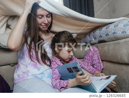 Mother and daughter sit in a hut made of a blanket and read a book. Pleasant time with children, quiet time before bed. Mother and daughter sit in a hut made of a blanket and read a book. Pleasant time with children, quiet time before bed. 126326685