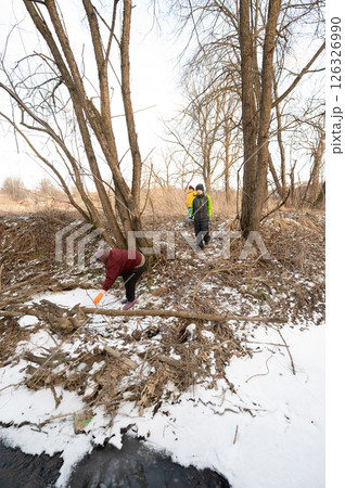 Exploring a snowy landscape by the river, individuals engaged in cleanup activities amidst winter foliage and bare trees under a clear sky 126326990