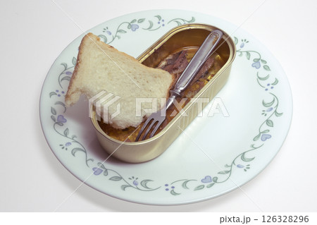 canned fish, sardines in oil, and small metal fork with three prongs. open tin can without lid and half of slice of toast bred, on porcelain plate. with clipping path 126328296