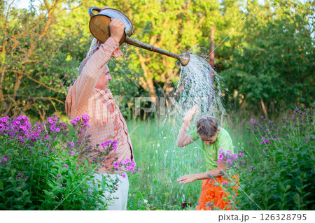 The father waters his son with water from a garden watering can, and the grandson rejoices and laughs. The father waters his son with water from a garden watering can, and the grandson rejoices and laughs. 126328795