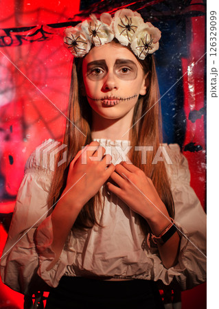 A teenage girl at a Halloween party with a skull painted on her face looks at the camera in fear. 126329099