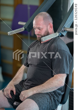 A man is sitting on a bench in a gym, wearing a black shirt and gray shorts. He is in a relaxed state, possibly meditating or taking a break from his workout 126329149