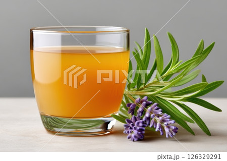 A clear glass of golden herbal tea accompanied by fresh lavender sprigs with vibrant purple blooms and green leaves. The image is captured against a neutral gray background on a light surface. 126329291