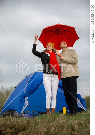An elderly couple stands under an umbrella near a tourist tent in the rain. Travel of an elderly man and woman in nature. An elderly couple stands under an umbrella near a tourist tent in the rain. Travel of an elderly man and woman in nature. 126329510