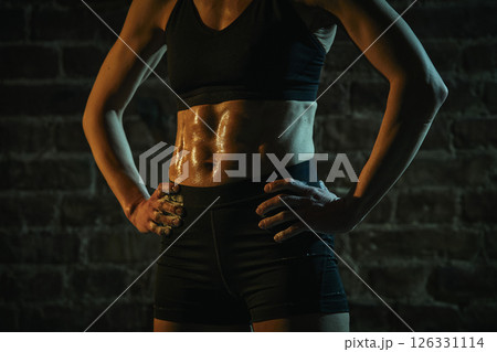 Cropped photo. Woman stands in gym with hands on hips and tight abs glistening with sweat, radiating strength, resilience and post-workout pride under dramatic light. Cropped photo. Woman stands in gym with hands on hips and tight abs glistening with sweat, radiating strength, resilience and post-workout pride under dramatic light. 126331114
