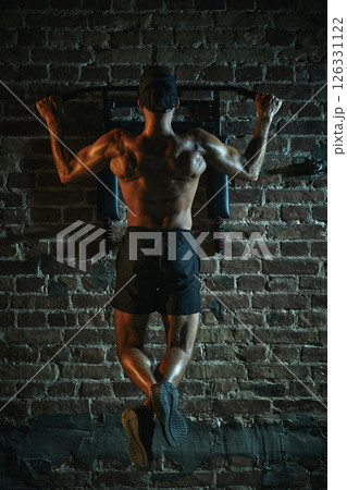 Rear view of man hanging on bar doing pull-up with muscular back, glistening under dramatic light, showcasing dedication and powerful physique. Rear view of man hanging on bar doing pull-up with muscular back, glistening under dramatic light, showcasing dedication and powerful physique. 126331122