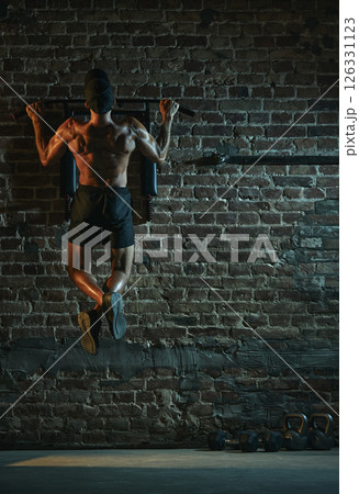 Rear view of muscular man performing pull-up from back view with defined upper body and crossed legs under spotlight in gritty gym atmosphere. 126331123