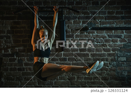 Woman lifting straight legs while hanging from bar in gym performing challenging core exercise with control, strength and balance. 126331124
