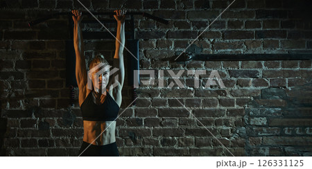 Focused woman gripping bar overhead ready for pull-up exercise, showing toned abdominal muscles and strong arms in dramatic gym lighting. Focused woman gripping bar overhead ready for pull-up exercise, showing toned abdominal muscles and strong arms in dramatic gym lighting. 126331125