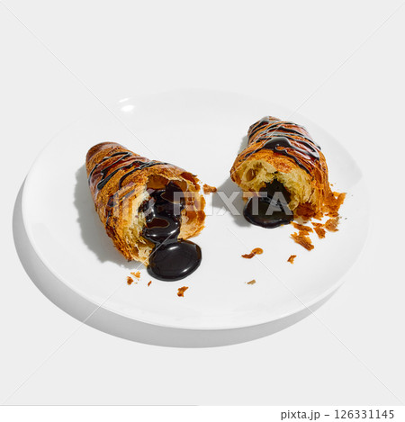 Chocolate-filled croissant sliced open on plate with glossy dark filling oozing onto surface, surrounded by golden crumbs against white background. Chocolate-filled croissant sliced open on plate with glossy dark filling oozing onto surface, surrounded by golden crumbs against white background. 126331145